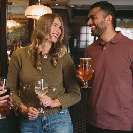 Three people standing at a pub bar holding a glass of red wine, a glass of white wine and a pint of beer.