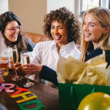 A group of friends sat in the interior seating area in a Greene King pub with drinks celebrating a birthday, A birthday banner is on the table with balloons and presents.