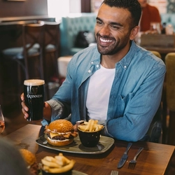 Person seated at a pub table holding a pint of Guinness, with a bacon cheeseburger and chips served in front.