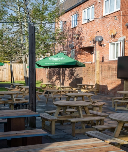 The exterior beer garden seating area at the Green Posts, with wooden picnic tables, shade umbrellas, a TV, and string lights overhead.