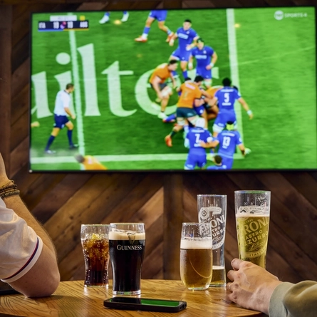 Two people sitting at a table in a pub, watching sports on the TV on the wall in front of them. A round of drinks and a mobile phone sit on the table between them.
