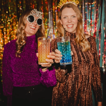 Two adults pose in front of a shimmering gold and silver tinsel backdrop, holding colourful cocktails toward the camera. One person wears novelty New Year-themed sunglasses and a glittery purple top, while the other wears a “Happy New Year” headband and a sparkling bronze dress. A bright blue cocktail and a peach-coloured cocktail are featured in the foreground, creating a festive New Year’s Eve party scene.