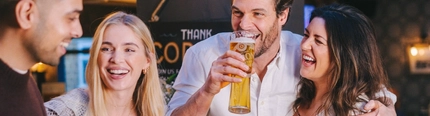 Four people standing inside a pub and laughing together, two of them holding glasses of beer, and two of them holding cocktails.