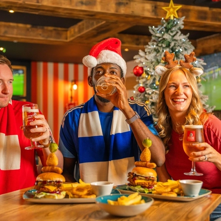 Three football fans sit around a wooden table in a sports pub decorated for Christmas, with a frosted Christmas tree and live football shown on large screens in the background. One person wears a Santa hat, another wears reindeer antlers, and team scarves add to the matchday atmosphere. Burgers topped with onion rings and pickle skewers, chips and glasses of beer are served on the table as the group celebrates during a festive Boxing Day football gathering.