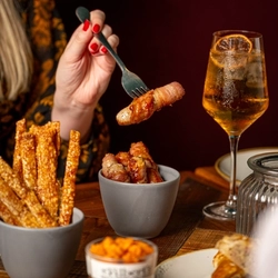 A person is holding a fork with a piece of Pigs in Blankets over a small gray bowl. 