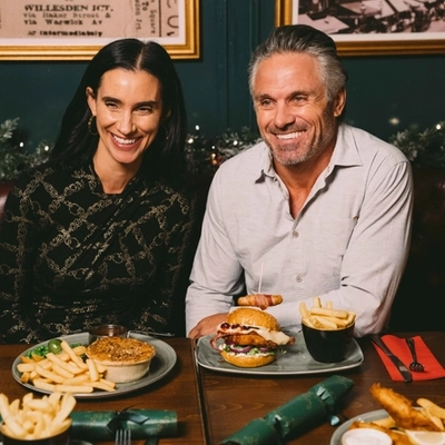 Two people sitting at a restaurant table with a plate of pie, chips, peas, and gravy; and a plate of a burger and chips in front of them. Christmas crackers, other plates of food, and glasses of Level Head, Hazy Day, and white wine also sit on the table; and a Christmas garland lit with fairy lights is visible in the background.