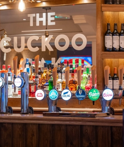 The bar and interior seating area at the Cuckoo Oak in Madeley, with bottles of wine on wooden shelves behind the counter, and illuminated beer taps.