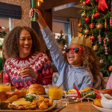 A festive dining table set for a Christmas meal beside a decorated Christmas tree with red and gold baubles, candy cane ornaments and warm fairy lights. Two children sit at the table, with one wearing a paper crown and red novelty sunglasses while holding a Christmas cracker overhead. An adult sits nearby holding a glass of sparkling drink. Plates of traditional festive food, glasses of orange juice, gravy boats, Christmas crackers and seasonal table decorations are spread across the table in a cosy, Christmas-themed restaurant setting.