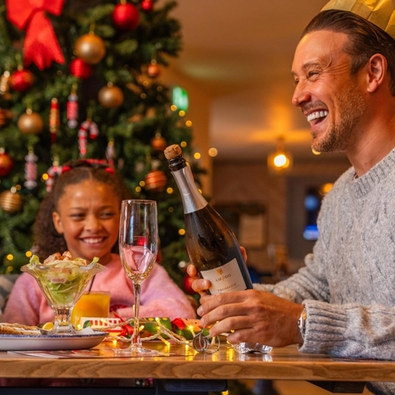 A festive dining table set beside a decorated Christmas tree with red and gold baubles, candy cane ornaments and warm fairy lights. One diner wearing a paper crown from a Christmas cracker holds a bottle of sparkling wine beside an empty flute glass, while two children sit nearby at the table, one wearing red novelty sunglasses. Plates of food, glasses of orange juice, Christmas crackers and string lights are arranged across the table, creating a warm Christmas restaurant setting.