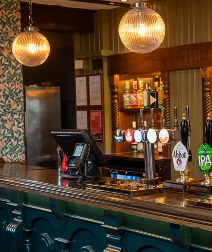 A close up view of the bar and beer taps inside The Hollybush in Lichfield, with golden drinks trays on the counter, and three spherical glass lights hanging above.
