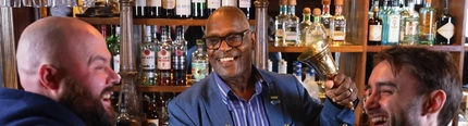 A man stood behind the bar in a Greene King venue holding a bell in his hands whilst 2 men stand at the bar holding pints of beer smiling. 