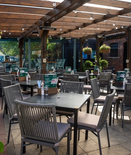 The paved, exterior beer garden seating area at The Ship in Bedford; with a large wooden pergola sheltering the tables and chairs, and flower baskets and string lights hanging from the dark wooden beams.