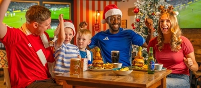 A group of adults and children sit around a wooden table in a sports pub decorated for Christmas, with a frosted Christmas tree and live football shown on large screens in the background. Two people wear Santa hats and another wears reindeer antlers as they share burgers, chips and drinks, including Guinness, Level Head beer, Heineken 0.0 and a children's juice carton. The festive family gathering combines Christmas dining with a football matchday atmosphere.
