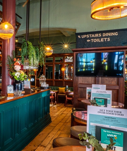 The wood panelled bar and interior restaurant seating area of The Fountains Abbey, with metal tables, stool seats, a TV on the wall, and plants hanging above the bar.