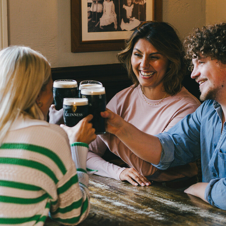 A group of people seated at an indoor wooden table, holding pints of Guinness in a Cheers pose and enjoying conversation.