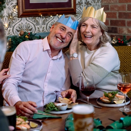 Two people seated together in a festively decorated pub wearing paper crowns, one blue and one gold. Plates of food, glasses of red and rosé wine, a pint of beer and green Christmas crackers are arranged on the table. Seasonal garlands, a Christmas wreath, patterned wallpaper and exposed brick walls create a festive dining setting.