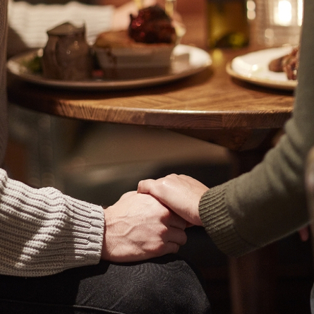A couple at a dark wood table holding hands. The image is taken from behind and it is cut off at the shoulders.