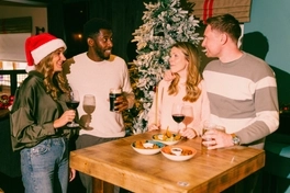 Four people gather around a high table in a pub decorated for Christmas, with a snow-covered Christmas tree in the background. The table is set with drinks including red wine, stout and lager, alongside sharing dishes and dipping sauces, creating a festive social dining scene.