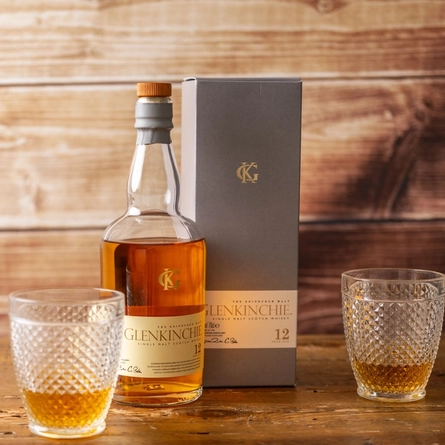 A bottle of March's Malt Of The Month - Glenkinchie whisky. The bottle stands on a wooden table with its presentation box, in between two glasses of the whisky.