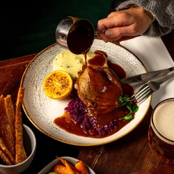 An overhead shot of a plated roast duck dish on a festive decorated wooden table, with the hands of a person not visible in the shot, pouring gravy from a mini pan over the plate.