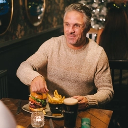 Two people sitting at a restaurant table, a burger served on a plate with a small bowl of fries and a plate of roast dinner sitting in front of them. A glass of Guinness and a Christmas cracker also sit on the table, and a Christmas tree is visible in the background.