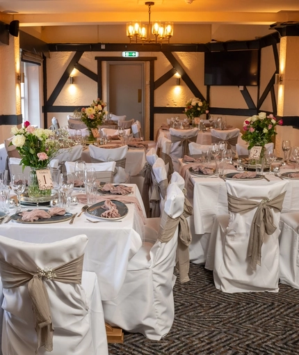 The function room at The Cherry Tree, set up for a wedding; with a TV on the wall, white tablecloths and seat covers, pink napkins, and vases of flowers on each table.