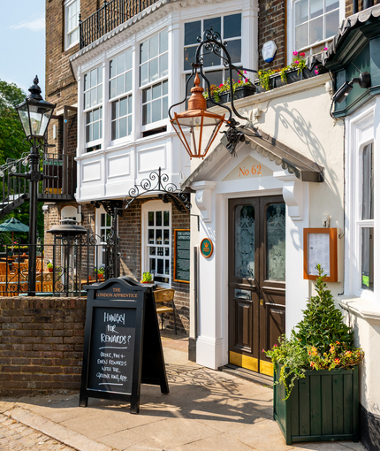 The exterior facade, main entrance, and beer garden seating area of The London Apprentice, with wooden tables, shade umbrellas, and string lights in the beer garden; a large golden lantern hanging above the double doors; and a green, wooden planter box filled with plants and flowers beside the entrance.