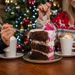 A close up view of a slice of Candy Cane Lane cake served on a plate which sits on a wooden table next to two mugs of hot drinks on saucers, two Christmas crackers, and a jug of cream. Two people are sitting at the table, holding their forks above the cake.