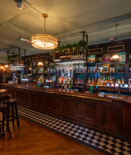 The wood panelled bar and interior restaurant seating area at The Plough in Bloomsbury, with wine glasses hanging in racks above the counter, and bottles of wine and alcoholic spirits on shelves behind the bar.