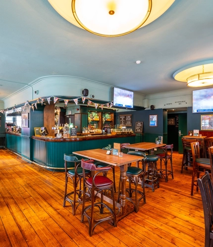 The bar and interior restaurant seating area at the Royal George in Euston; with wooden flooring, an upholstered booth seat, TVs on the walls, and gaming machines.