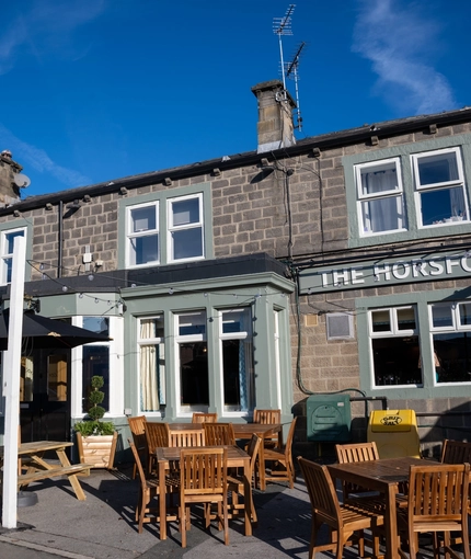 The exterior facade, signage, and seating area at The Horsforth in Horsforth, with wooden picnic tables, shade umbrellas, and string lights over the seating area.