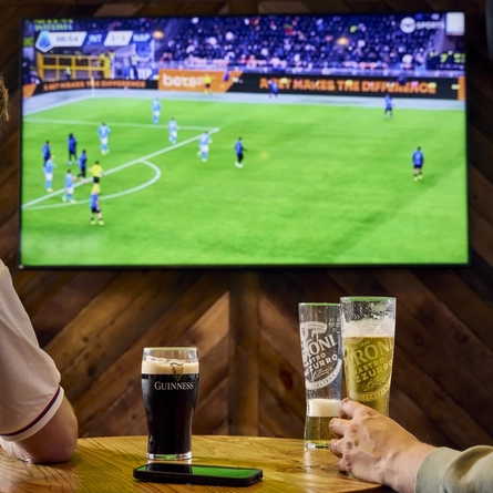 Two people sitting at a table in a pub, watching sports on the TV on the wall in front of them. A glass of Guinness, two glasses of Peroni, and a mobile phone sit on the table.
