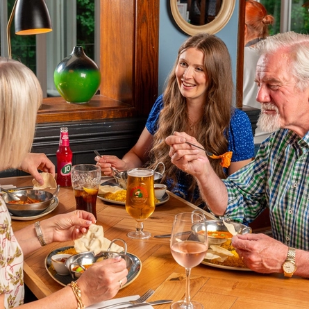 A group of four people sitting at a wooden restaurant table, enjoying dishes of curry and rice served with poppadoms. Glasses of wine, lager, Coca-Cola and a bottle of J2O sit on the table in front of them.