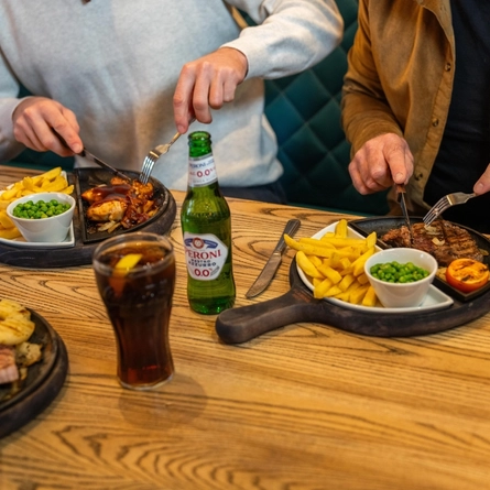 A close up view of two plates of food, a glass of fizzy drink, and a bottle of Peroni 0.0% alcohol beer, sitting on a wooden restaurant table. Two people sitting at the table are cutting up the food with their knives and forks.