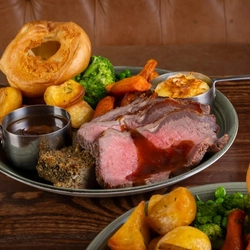 Two plates of roast dinner, including meat, Yorkshire pudding, vegetables and gravy, sitting on a wooden table.