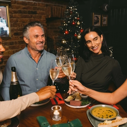 A close up view of four people sitting at a restaurant table laden with plates of food, clinking together glasses of Lanson champagne. Christmas crackers and the bottle of champagne sit on the table, and a Christmas tree decorated in red and gold is visible in the background.