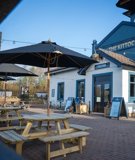 The exterior facade, signage, and beer garden seating area of The Kittoch, with wooden picnic tables, shade umbrellas, and string lights overhead.
