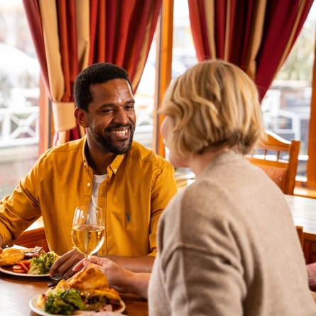Two people sitting at a restaurant table with plates of roast dinner in front of them. One person is holding a glass of wine, the other is holding a glass of beer, and menus sit on the table along with salt and pepper.