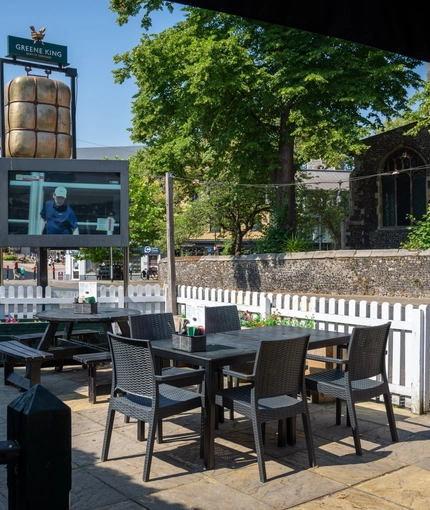 The exterior beer garden seating area of The Woolpack Inn; with wooden picnic tables, a TV, and a view of the surrounding buildings.