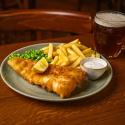 A close up view of Fish & Chips served on a plate with peas, a slice of lemon, and a small pot of dipping sauce. The plate sits on a wooden restaurant table along with a glass of beer.