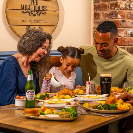 Three people seated around a wooden table in a pub, sharing Sunday roast meals. One person pours gravy from a small jug onto a plate of roast dinner with Yorkshire puddings, vegetables and potatoes. A bottle of alcohol-free beer, a pint of Guinness and a children's juice carton sit among the dishes. The background features blue panelled walls, exposed brickwork and a wooden barrel-style sign reading “The Mill House”.