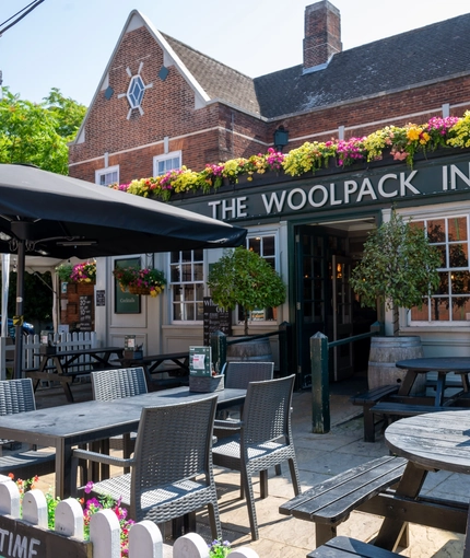 The exterior facade, signage, and beer garden seating area at The Woolpack Inn, with wooden picnic tables, trees on each side of the entrance, and a large shade umbrella.