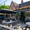 The exterior facade, signage, and beer garden seating area at The Woolpack Inn, with wooden picnic tables, trees on each side of the entrance, and a large shade umbrella.
