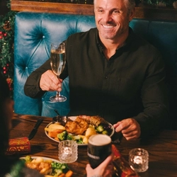 A person sitting at a restaurant table and raising a glass of champagne. A roast dinner served on a plate sits on the table in front of them, along with Christmas crackers and another plate of food.