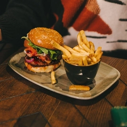 A festive decorated wooden table holding a plated burger and fries.  There is the torso of a woman holding a glass of sparkling wine sat at the table.