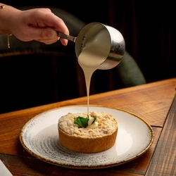 A hand pouring custard from a mini pan, over a plated apple crumble. on a festive decorated wooden table.