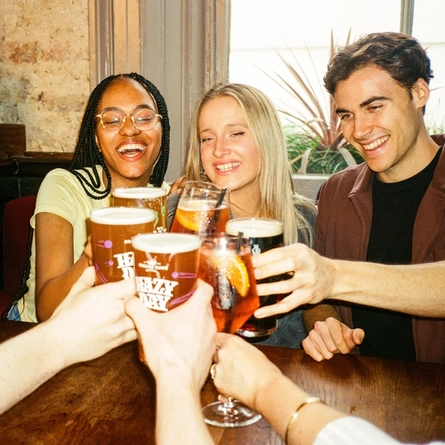 A group off friends sat in the interior restaurant and seating area in a Greene King venue in London holding drinks into a 'cheers' position.