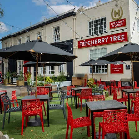 The exterior facade, signage, and beer garden seating area at The Cherry Tree; with shade umbrellas on some of the tables, and string lights overhead.