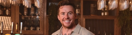 Danny Jones stood next to a bar, smiling at the camera whilst holding a pint of Level Head Ale.