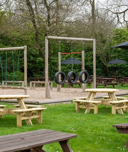 The exterior beer garden seating area and children's play area at The Hussey Arms in Brownhills, with wooden picnic tables, shade umbrellas, and obstacle course.